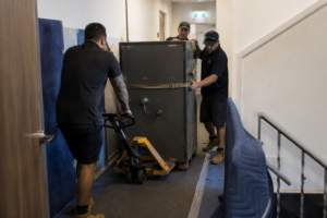 Professional movers transporting a heavy commercial safe through a narrow corridor using a pallet jack and protective wall coverings